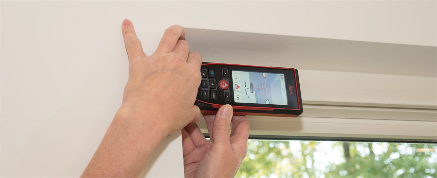 Two hands holding a Leica DISTO against the head rail of a window to take a measurement.